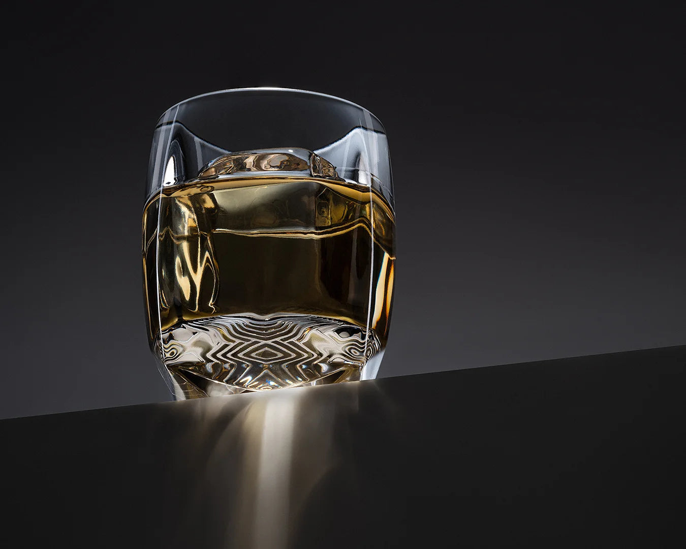 The Rauk Heavy Tumbler 250 mL Sinder (Grey) with whiskey sitting on the edge of a slate black ledge of a table, illuminated with an overhead light which casts a light pattern on the edge of the table.
